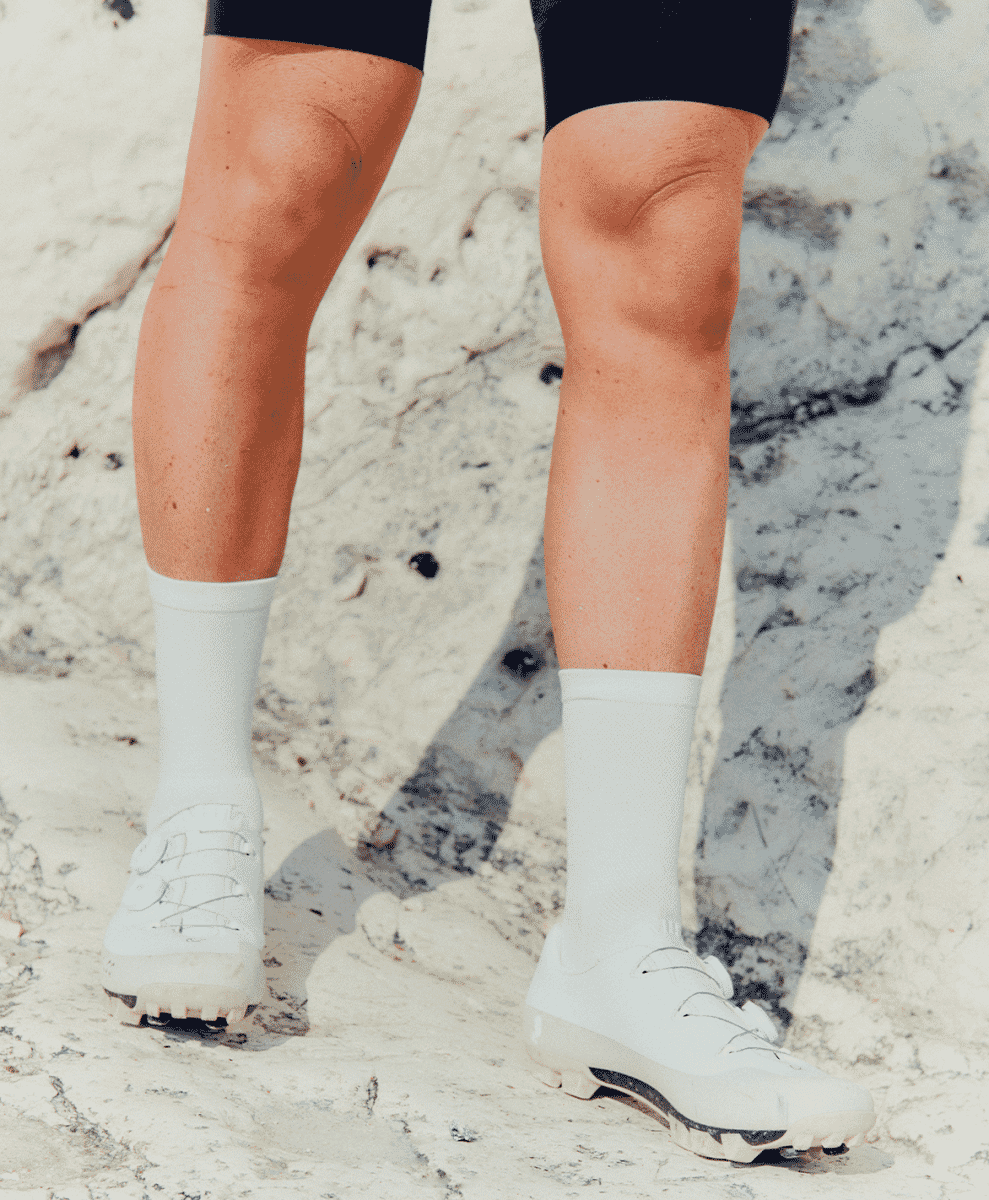 A cyclist's legs and white shoes standing on a rocky, textured surface under bright sunlight.