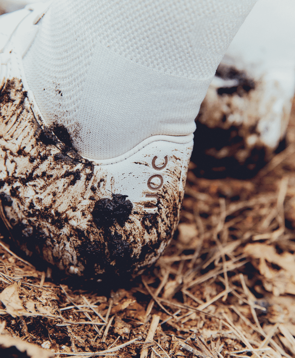 A mud-covered white QUOC cycling shoe against a rustic background of straw and dirt.