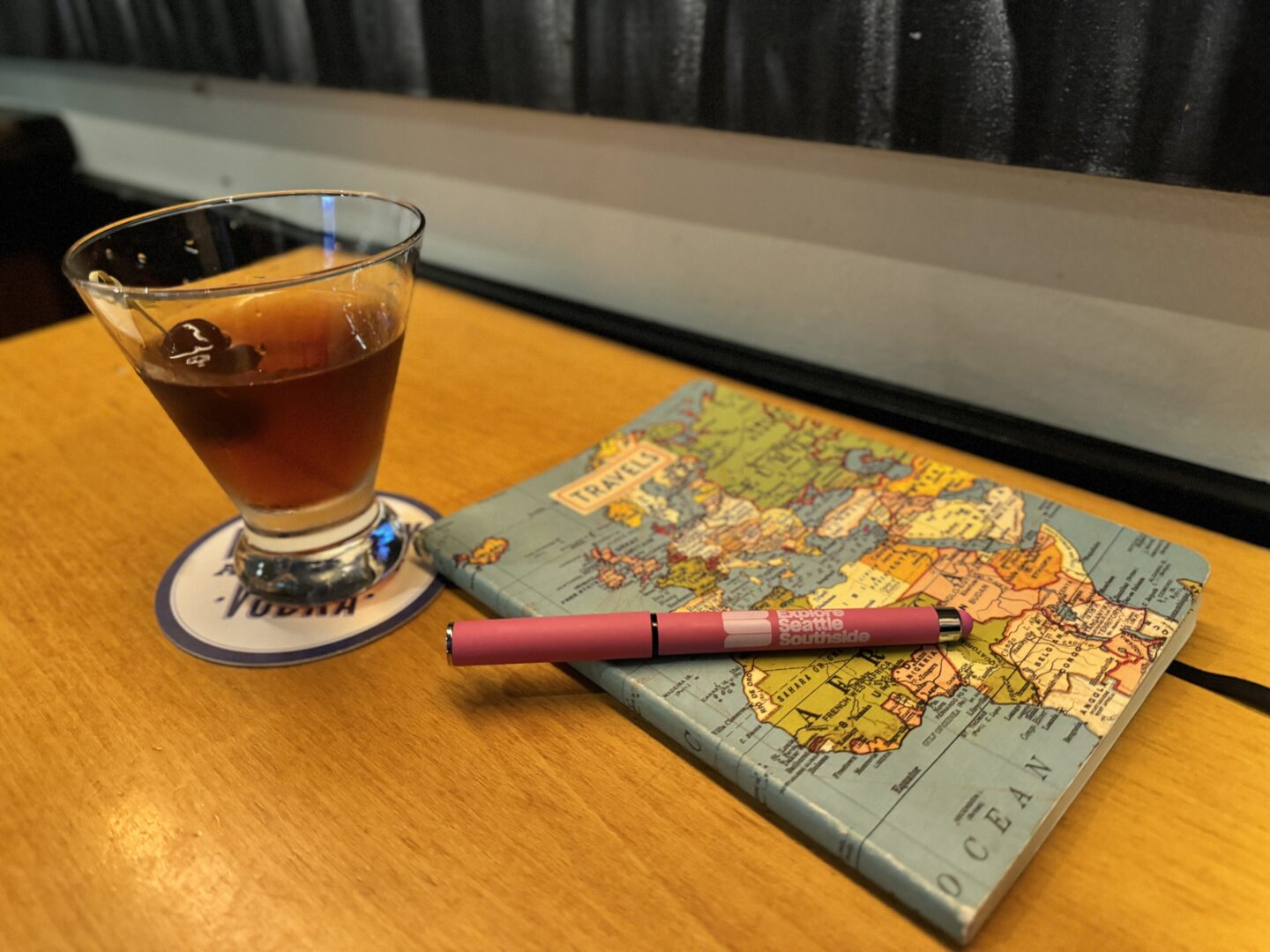 A Manhattan cocktail and a leather-bound travel journal on a restaurant table.