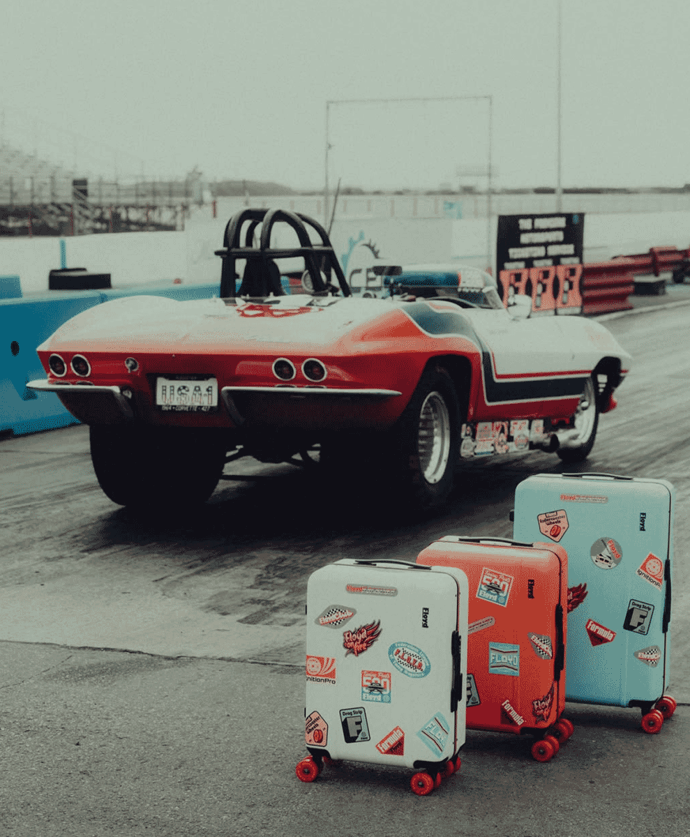 Three sticker-covered colorful suitcases in front of a vintage red and white race car.