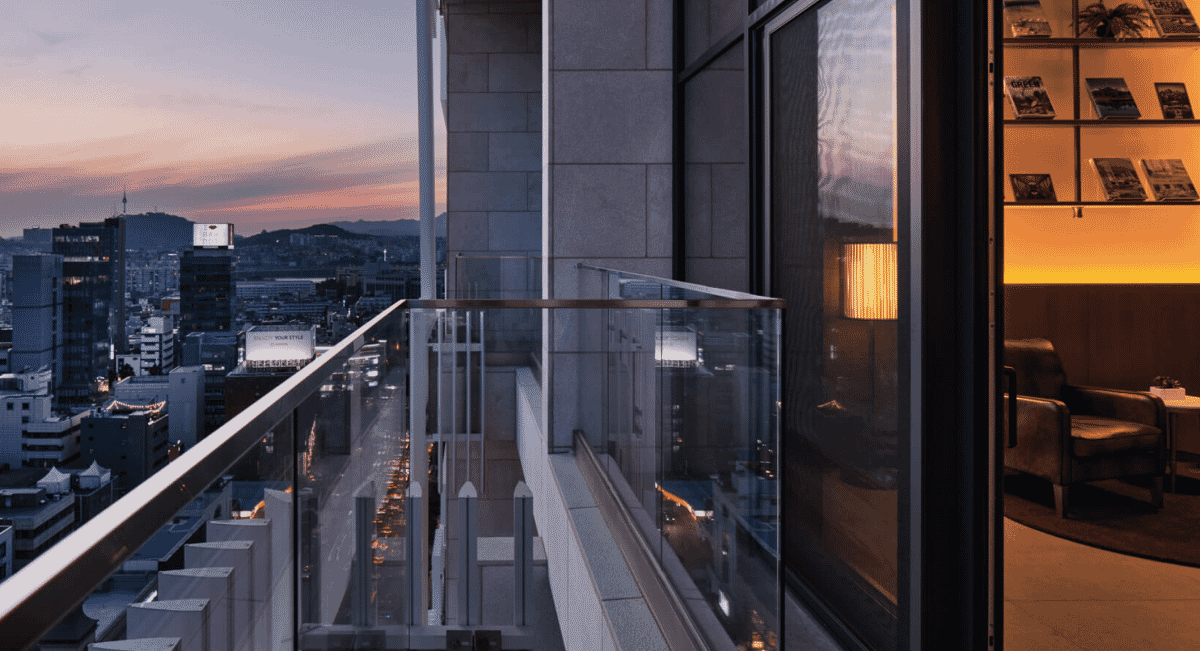 Evening cityscape viewed from a modern glass balcony with warm interior lighting visible through an open door.