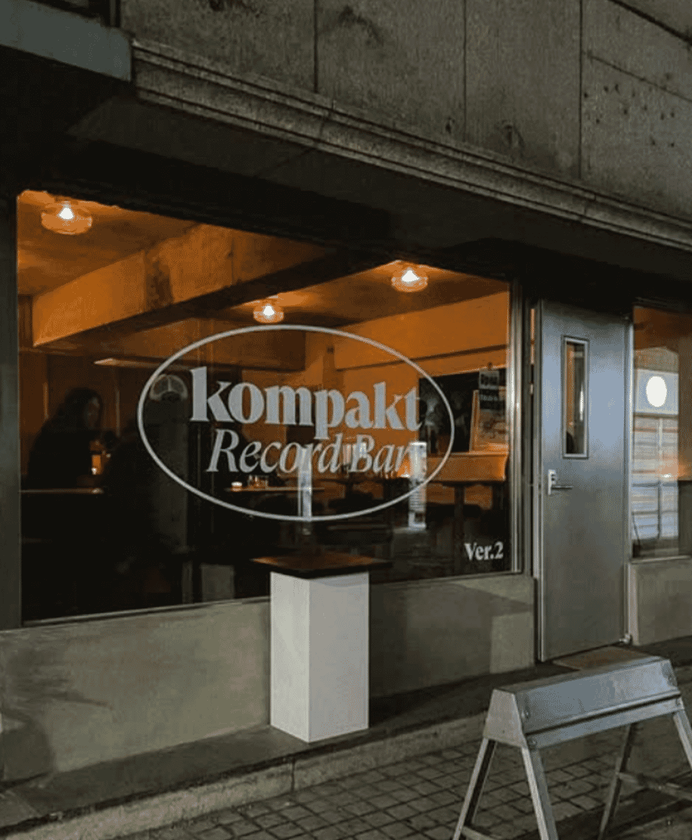 Exterior view of 'Kompakt Record Bar' at night, showing a warm-lit interior through a glass window.