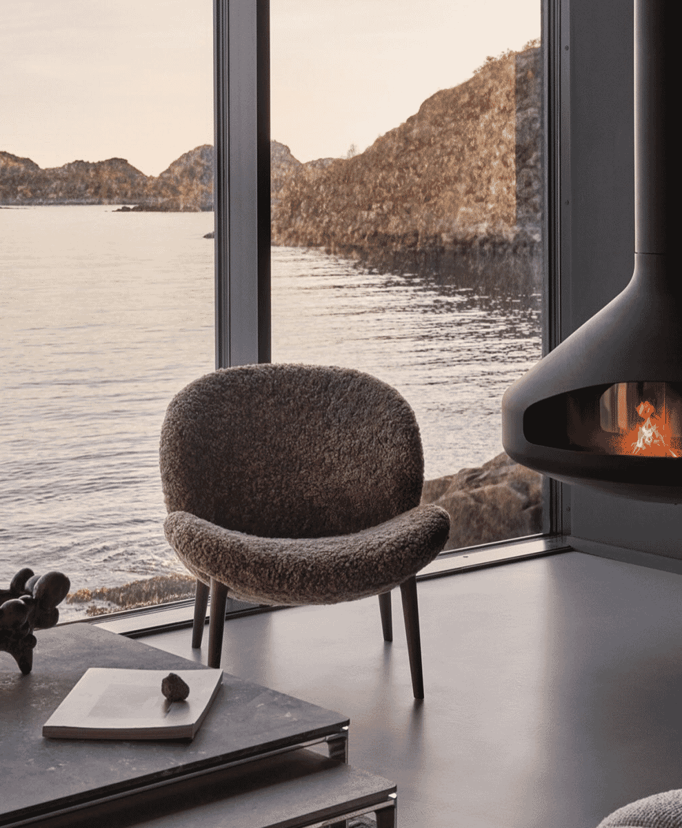A cozy interior featuring a textured wool armchair next to a fireplace with a coastal window view.
