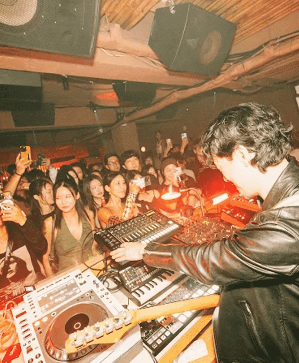 A DJ in a black leather jacket performing at a club under vibrant red lighting with an energetic crowd.