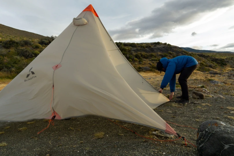 A gear tester in high-altitude clothing bracing a tent against extreme 50 mph wind gusts in Patagonia.